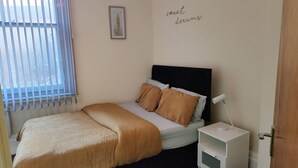 Une chambre à coucher avec un lit, une table de chevet, une lampe et un mur orné d’un tableau encadré et d’une inscription.