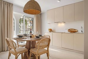 A dining area with a round table, four wicker chairs, a hanging light fixture, and a view of a cityscape through a window with decorative ironwork. - Villa Violeta Estepona Old Town (Estepona)