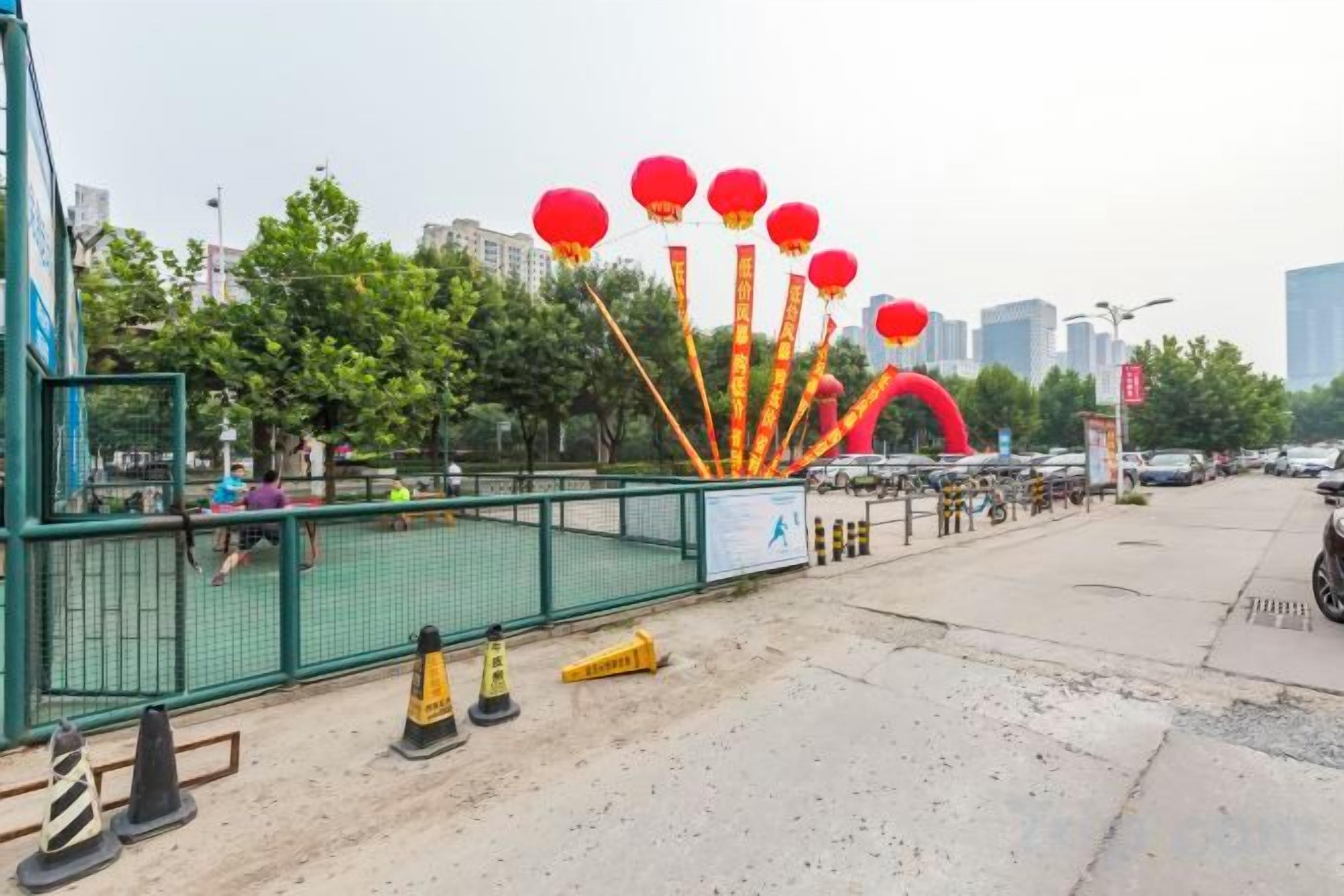 Un espace délimité par une clôture et une barrière verte, un terrain de basketball et une rangée de voitures garées.