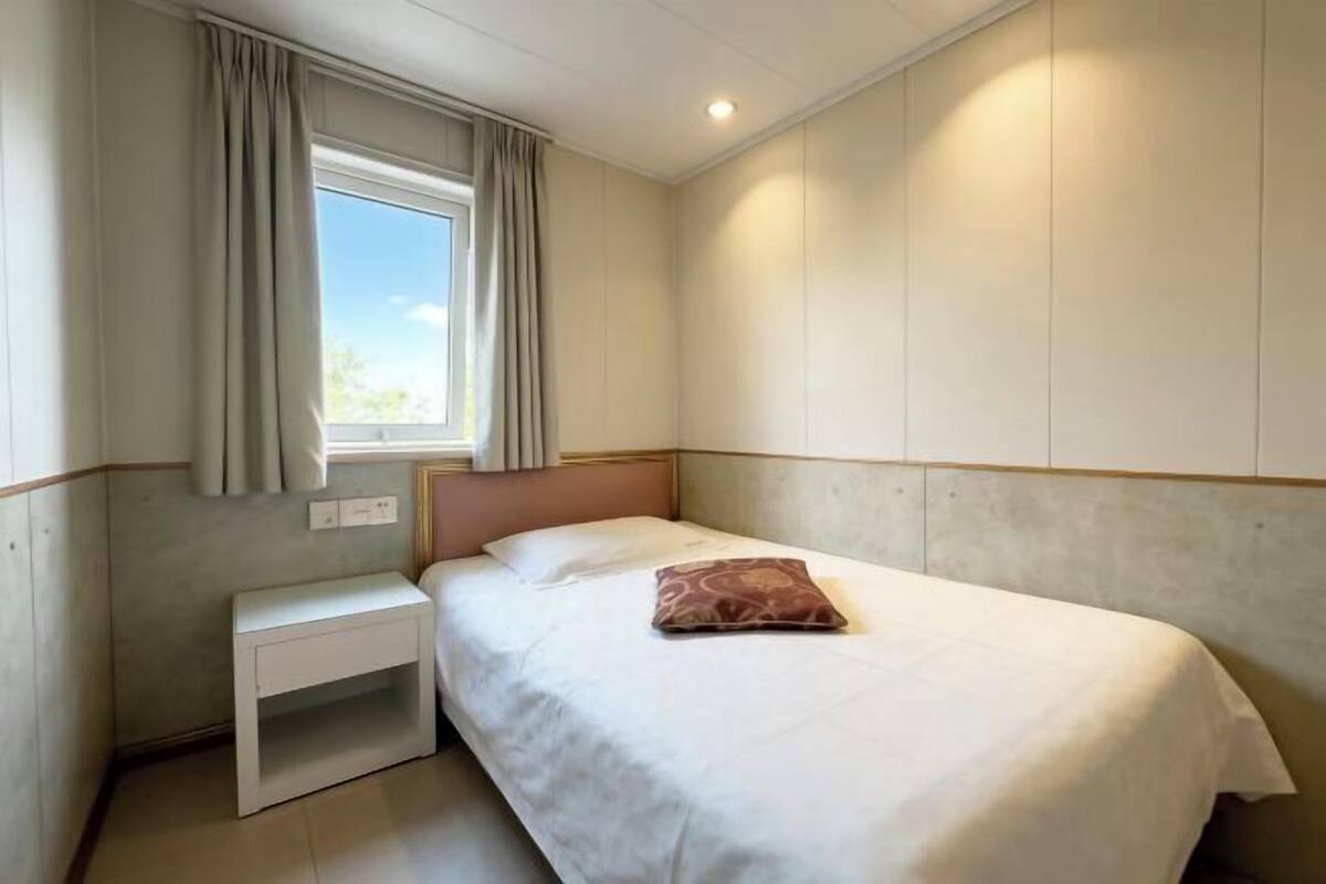 A bedroom with a bed, a nightstand, a window with curtains, and a wall-mounted light.