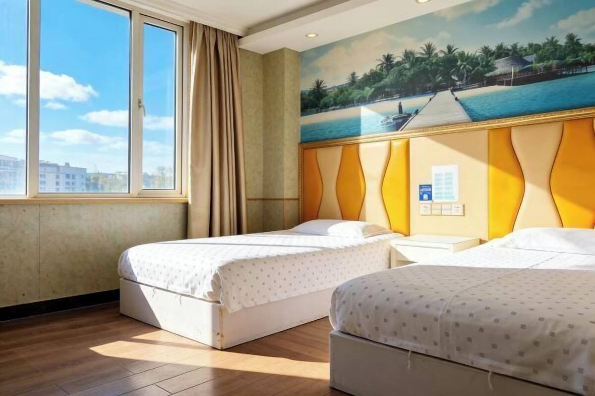 A hotel room with two beds, a large window, a painting of a beach, and a view of buildings outside.