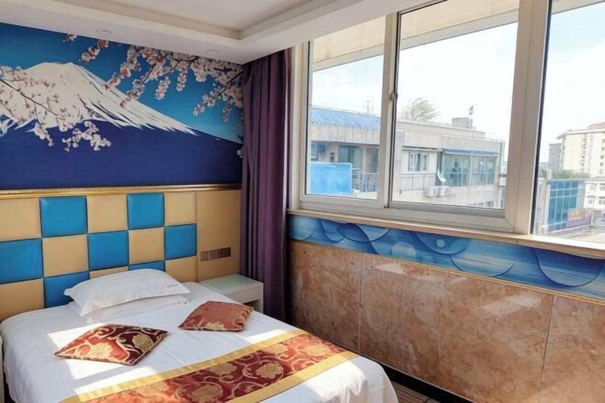 A hotel room with a bed, a mural on the wall, a window with a view of buildings, and a window blind.