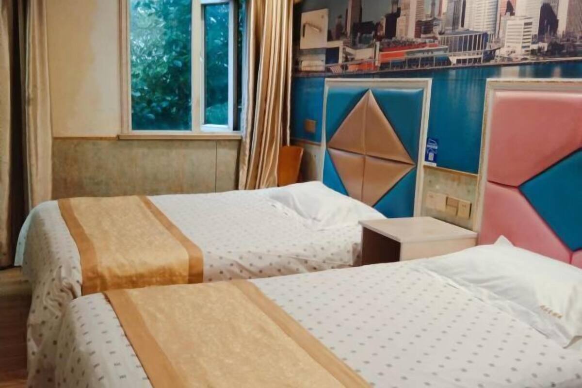 A hotel room with two beds, a window with curtains, a cityscape mural on the wall, and a small table.