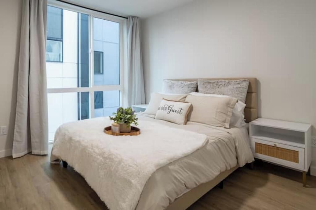 A bedroom with a large window, a bed with a white duvet, a bedside table, and a small plant on the bed.