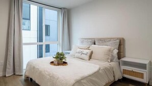 A bedroom with a large window, a bed with a white duvet, a bedside table, and a small plant on the bed. - Capitol Chill - Modern Luxury Townhouse (Seattle)