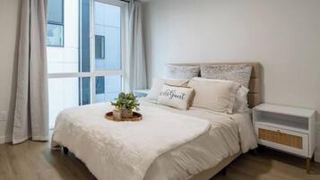 A bedroom with a large window, a bed with a white duvet, a bedside table, and a small plant on the bed.