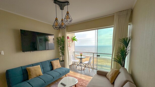 A modern living room with a sofa, a coffee table, and a TV mounted on the wall. There is a balcony with a view of the ocean.