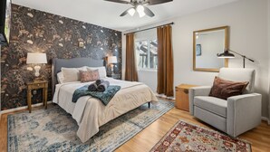 Ein Schlafzimmer mit einem Bett, einem Sessel, einem Deckenventilator, einem Fenster mit Vorhängen und einer Wand mit Blumenmuster.