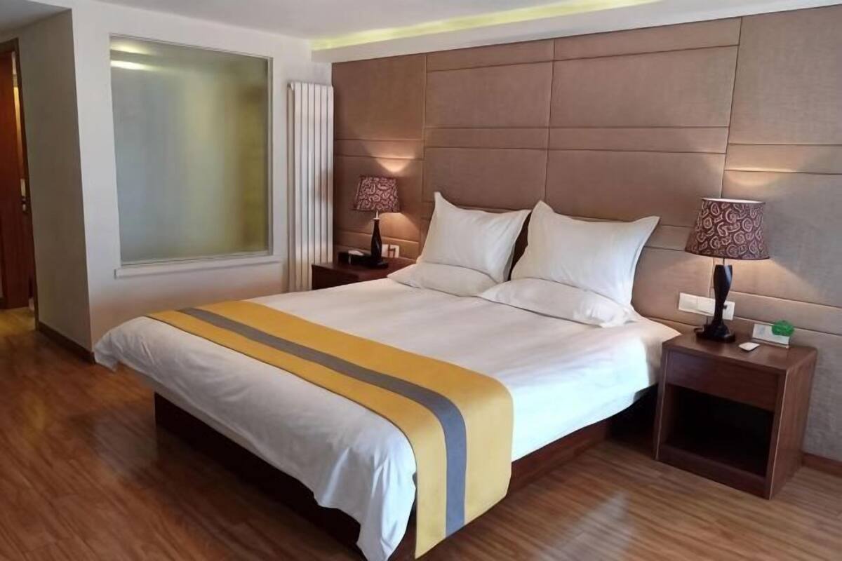 A hotel room with a large bed, two bedside tables with lamps, a window with blinds, and a wooden floor.