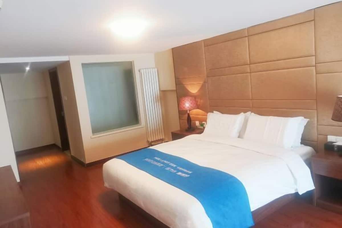 A hotel room with a large bed, a wooden headboard, a bedside lamp, and a window with blinds.