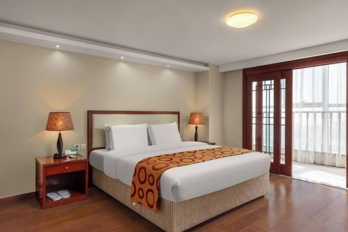 A hotel room with a large bed, two bedside lamps, a nightstand with a shoe rack, and a view of the outdoors through a large window.