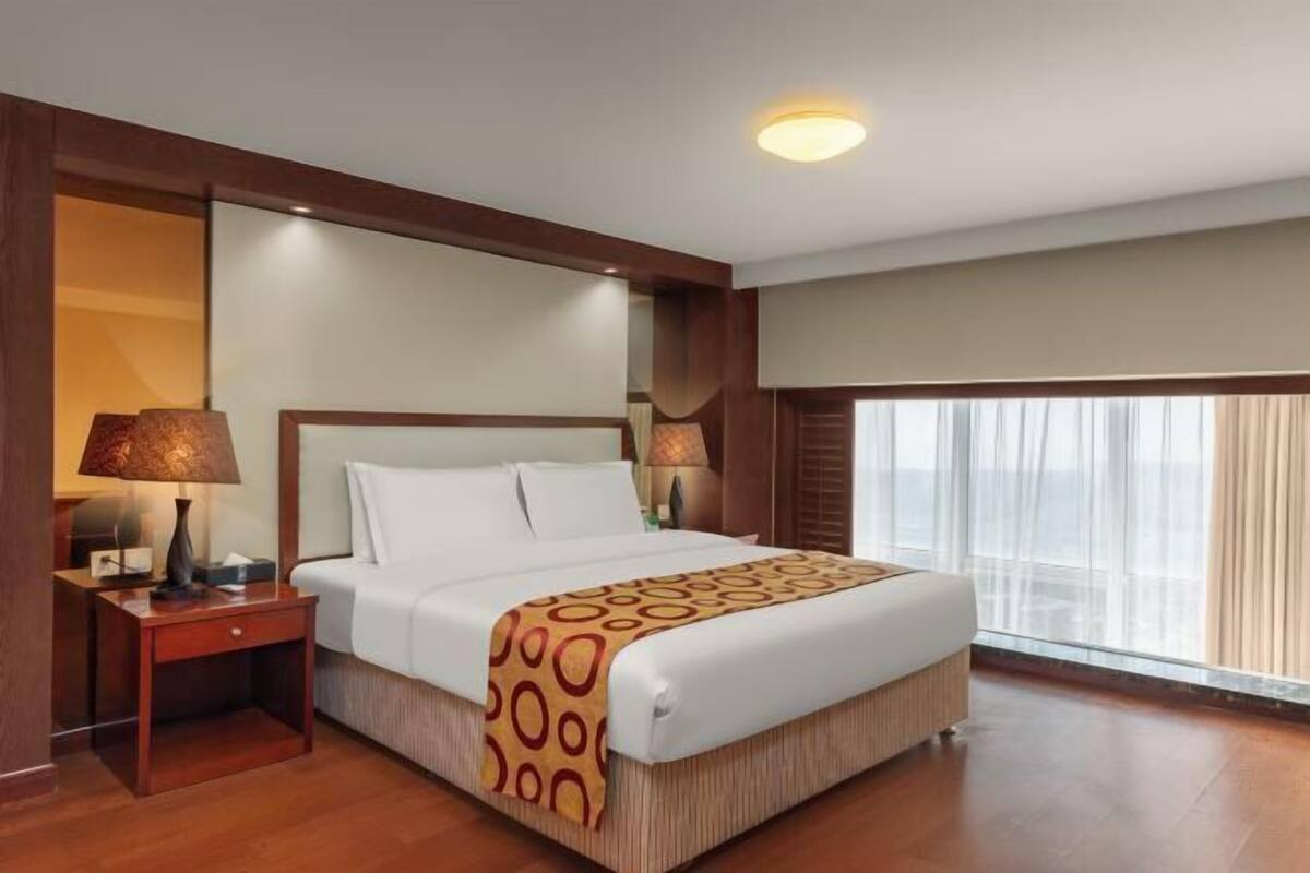 A hotel room with a large bed, two bedside tables, a lamp, a window with curtains, and a ceiling light.