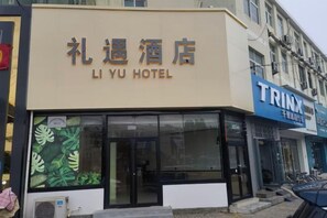 Un hotel llamado 'Li Yu Hotel' con un gran letrero sobre la entrada, una unidad de aire acondicionado visible y una tienda con un cartel azul a la derecha.