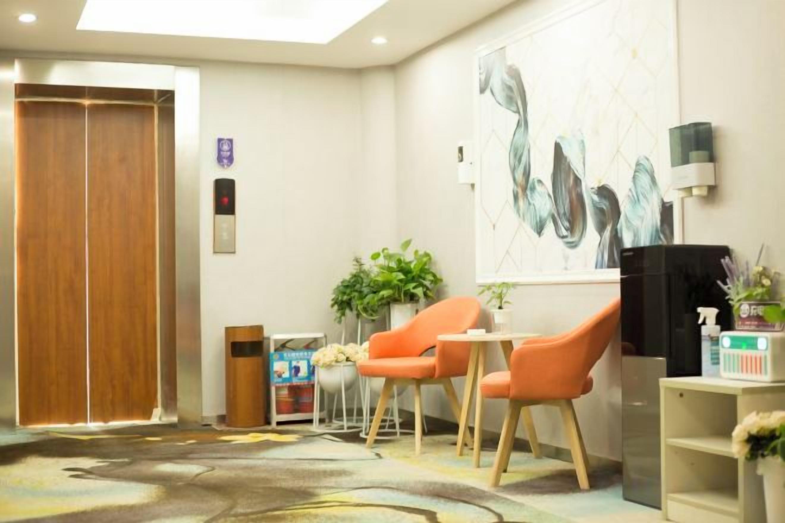 A hotel lobby with a reception desk, two orange chairs, a small table, a potted plant, a poster, and an elevator.