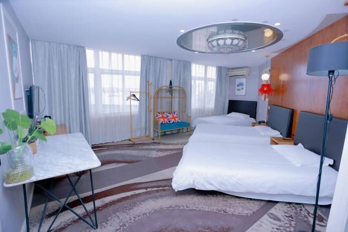 Ein Hotelzimmer mit zwei Betten, einem kleinen Tisch mit Pflanze, einem Vogelkäfig und einem großen Fenster mit Vorhängen.