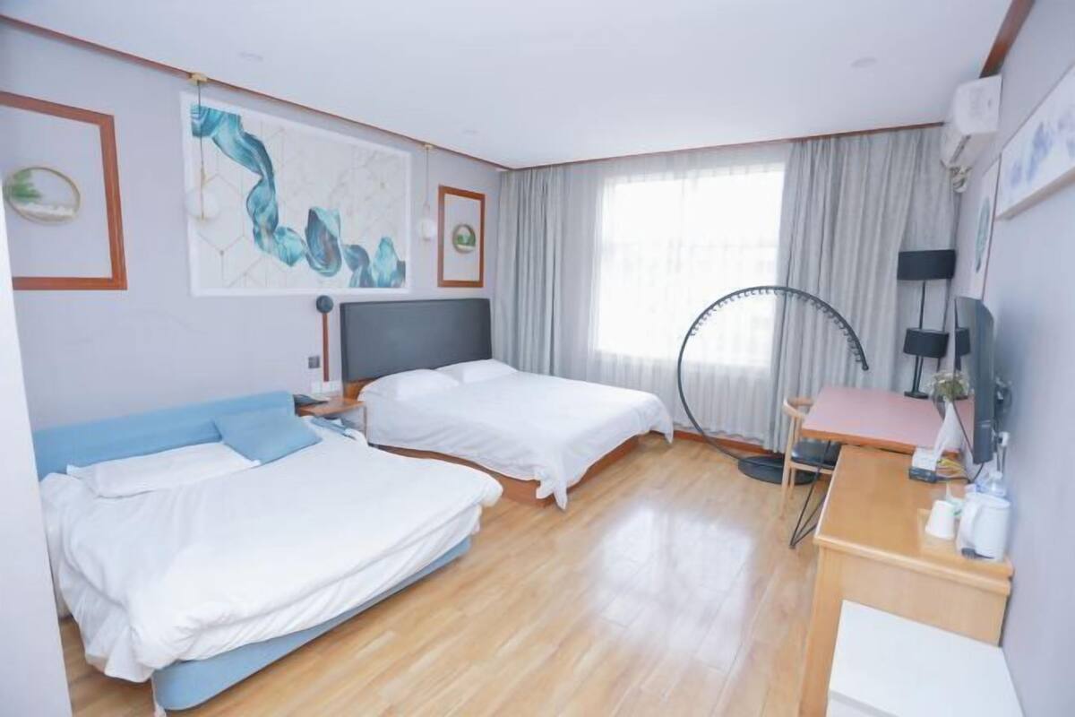 Ein Hotelzimmer mit zwei Betten, einem Schreibtisch, einem Stuhl, einem großen Fenster mit Vorhängen und Bildern an den Wänden.