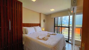 A hotel room with a large bed, a wooden wardrobe, a bedside table with a lamp, and a window with a view of the beach. - Flat Oceânica Frente Mar - By Almare (Joao Pessoa)