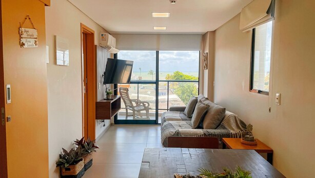 A modern living room with a sofa, a coffee table, and a TV. There is a balcony with a view of the beach and palm trees. - Flat Oceânica Frente Mar - By Almare (Joao Pessoa)