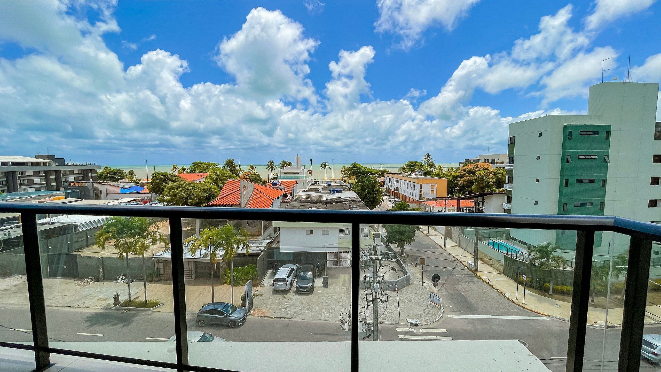 A view from a balcony overlooking a street with cars, buildings, and a beach in the distance.