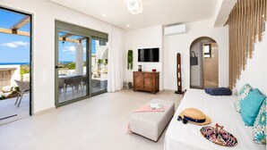 A modern living room with a glass door leading to a balcony, a flat-screen TV, a wooden cabinet, and a sofa with a hat and sunglasses on it.