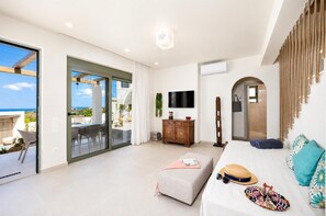 Un salon moderne avec une porte vitrée donnant sur un balcon, un téléviseur à écran plat, un meuble en bois et un canapé sur lequel sont posés un chapeau et des lunettes de soleil.