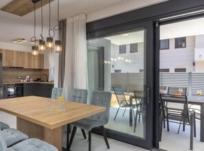 Une cuisine moderne avec des meubles en bois, un coin repas avec une table en bois et des chaises à revêtement gris, et une porte-fenêtre coulissante donnant sur un espace extérieur aménagé avec une table et des chaises.