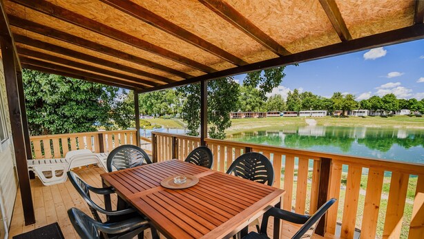 Un espacio al aire libre cubierto con una mesa y sillas de madera, vista a un lago y una hilera de vehículos recreativos al fondo.