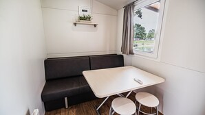 Ein kompakter Raum mit einem dunkelgrauen Sofa, einem kleinen Tisch und zwei Hocker. Es gibt ein Fenster mit Vorhängen, ein Regal mit einer Pflanze und eine Heizung an der Wand.