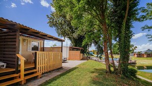 Eine Holzhütte mit einer Holzterrasse und einer kleinen Veranda, umgeben von einer Rasenfläche und einem Teich.