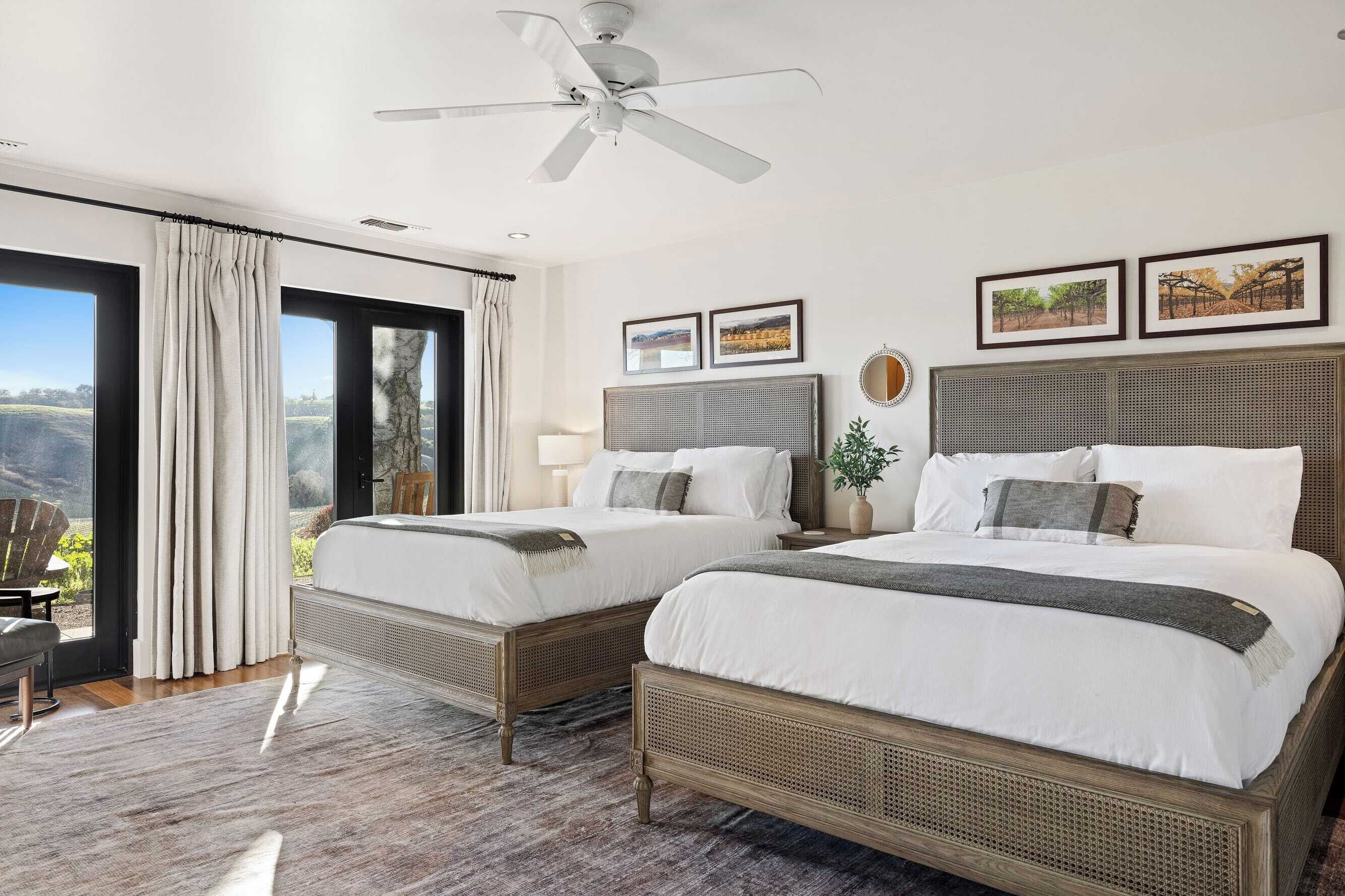 A hotel room with two beds, a ceiling fan, large windows with curtains, and framed pictures on the wall.