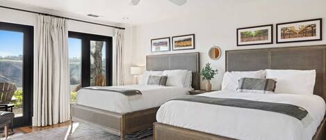A hotel room with two beds, a ceiling fan, large windows with curtains, and framed pictures on the wall.
