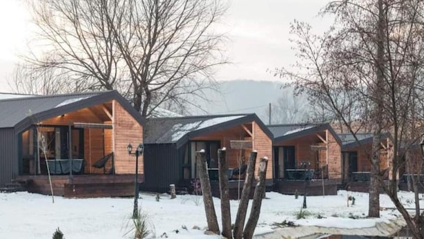 A row of cabins with wooden exteriors and large windows, situated in a snowy landscape with a calm body of water in the foreground.