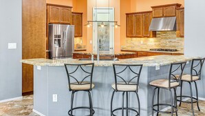 A kitchen with a central island, bar stools, wooden cabinets, a stainless steel refrigerator, and a stone backsplash.