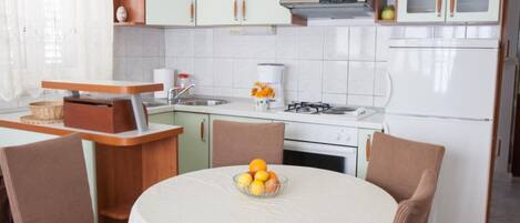 A kitchen with a round table, chairs, a bowl of fruit, a stove, and a refrigerator.