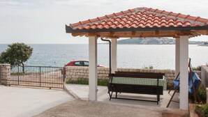 A covered outdoor seating area with a bench, a red car parked nearby, and a view of the sea. - One-Bedroom Apartment With Sea View Ap 2 (Sevid)