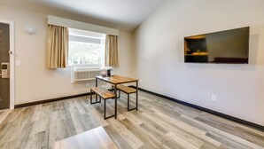 Ein Zimmer mit Holzboden, einem Tisch mit Bänken, einem Fenster mit Vorhängen und einem an der Wand befestigten Fernseher.