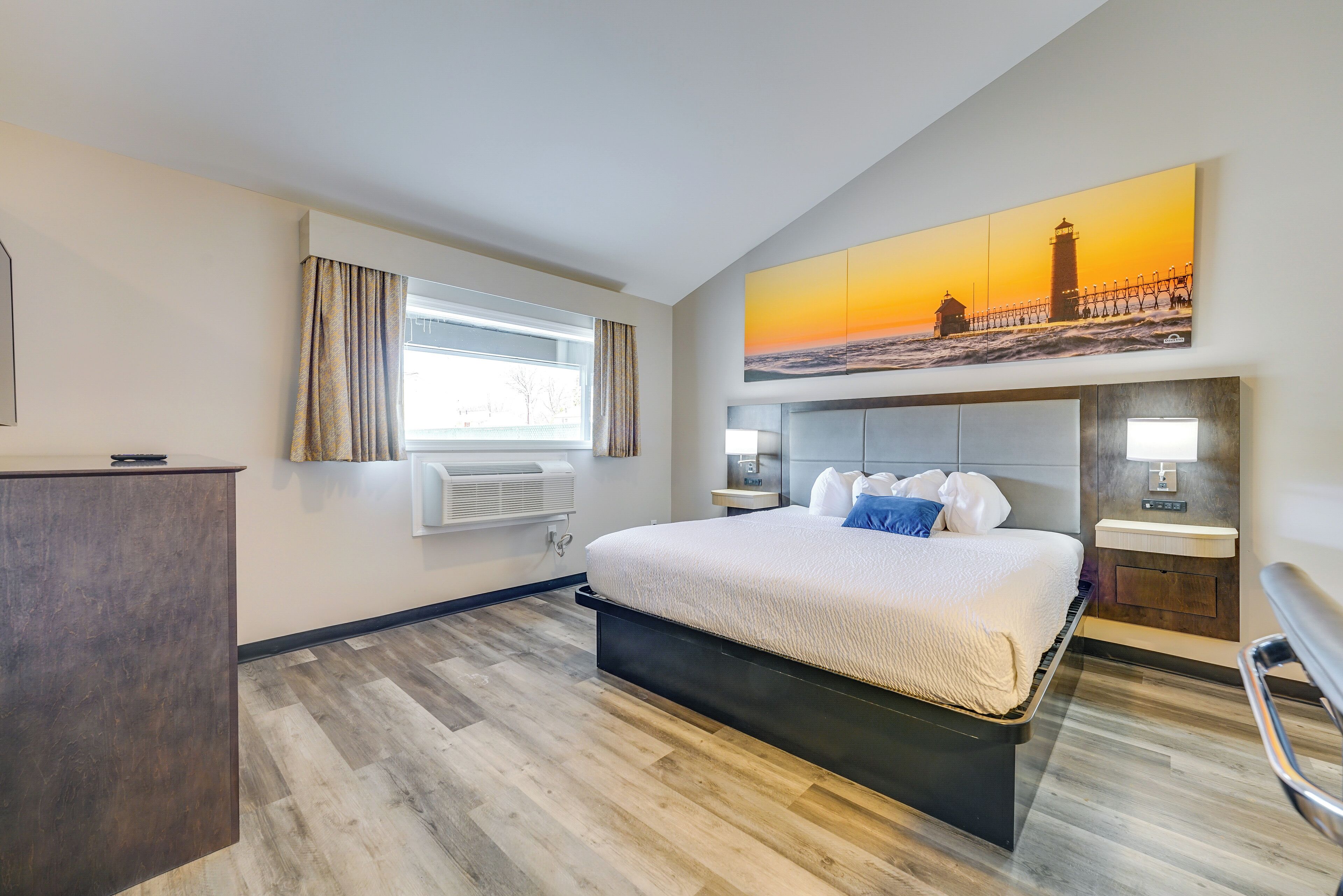 Een moderne hotelkamer met een groot bed, een bureau, een raam met gordijnen, een aan de muur bevestigd kunstwerk van een vuurtoren en een ingebouwde nachtkastje.