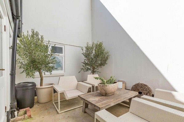A small, enclosed outdoor space with two potted olive trees, a wooden table, and a wicker sofa.