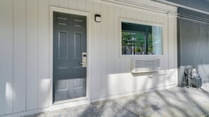 Fachada blanca con puerta gris, ventana con respiradero y calentador de agua pequeño al aire libre.