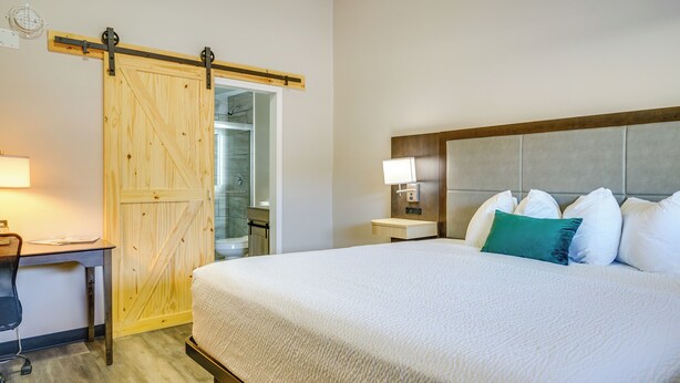Habitación de hotel con cama, escritorio, silla, mesita de noche, lámpara y puerta corredera al baño.