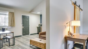 Un salón moderno con una mesa de centro de madera, un sofá estampado y un escritorio con lámpara. Se ve una puerta y un microondas al fondo.