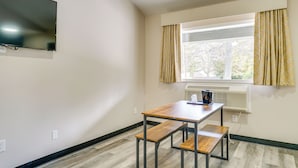 Una habitación pequeña y ordenada, con una mesa de madera y un banco, un televisor de pantalla plana en la pared y un ventanal con cortinas.