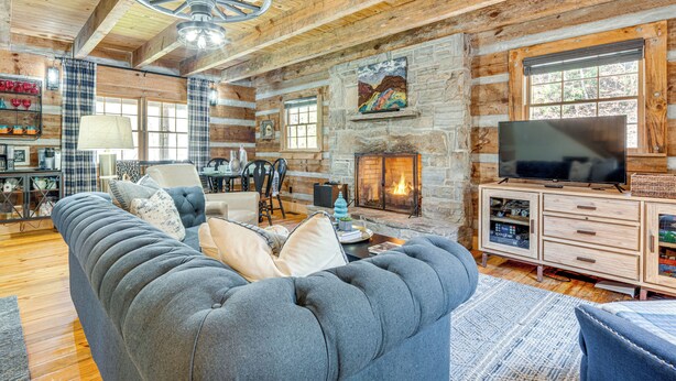 A rustic log cabin interior with a large sofa, fireplace, stone walls, and a television. - Hot Tub, Fire Pit & Creek Access: Whittier Cabin! (Whittier)
