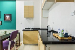 Une cuisine compacte avec un plan de travail noir, des placards en bois et un petit coin repas agrémenté d’une chaise violette.