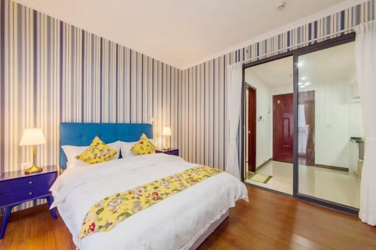 A bedroom with a striped wall, a blue headboard, a bed with white bedding and a floral throw, two bedside lamps, and a sliding glass door leading to a bathroom.