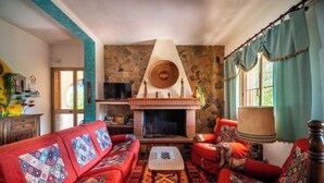 A cozy living room with a fireplace, red sofas, patterned cushions, and a stone wall. - Resonance Experience, Villa Retreat & Sound Healing (Sardegna)