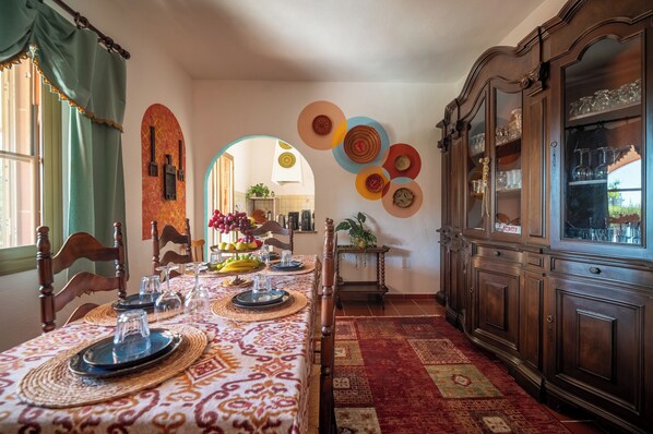 A dining room with a long table set for a meal, wooden chairs, a patterned tablecloth, and a red carpet.