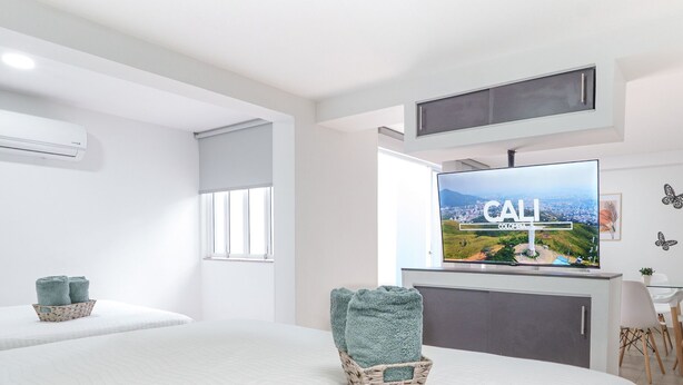 A hotel room with a bed, a television displaying 'Cali Colombia', a window, and a wall-mounted air conditioner. - Casa Saza (Cali)