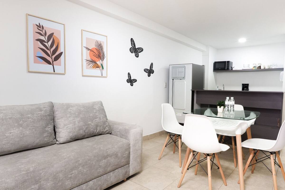 A modern living room with a grey sofa, white chairs, and a glass dining table. There are framed artworks on the wall and a black decorative butterfly.