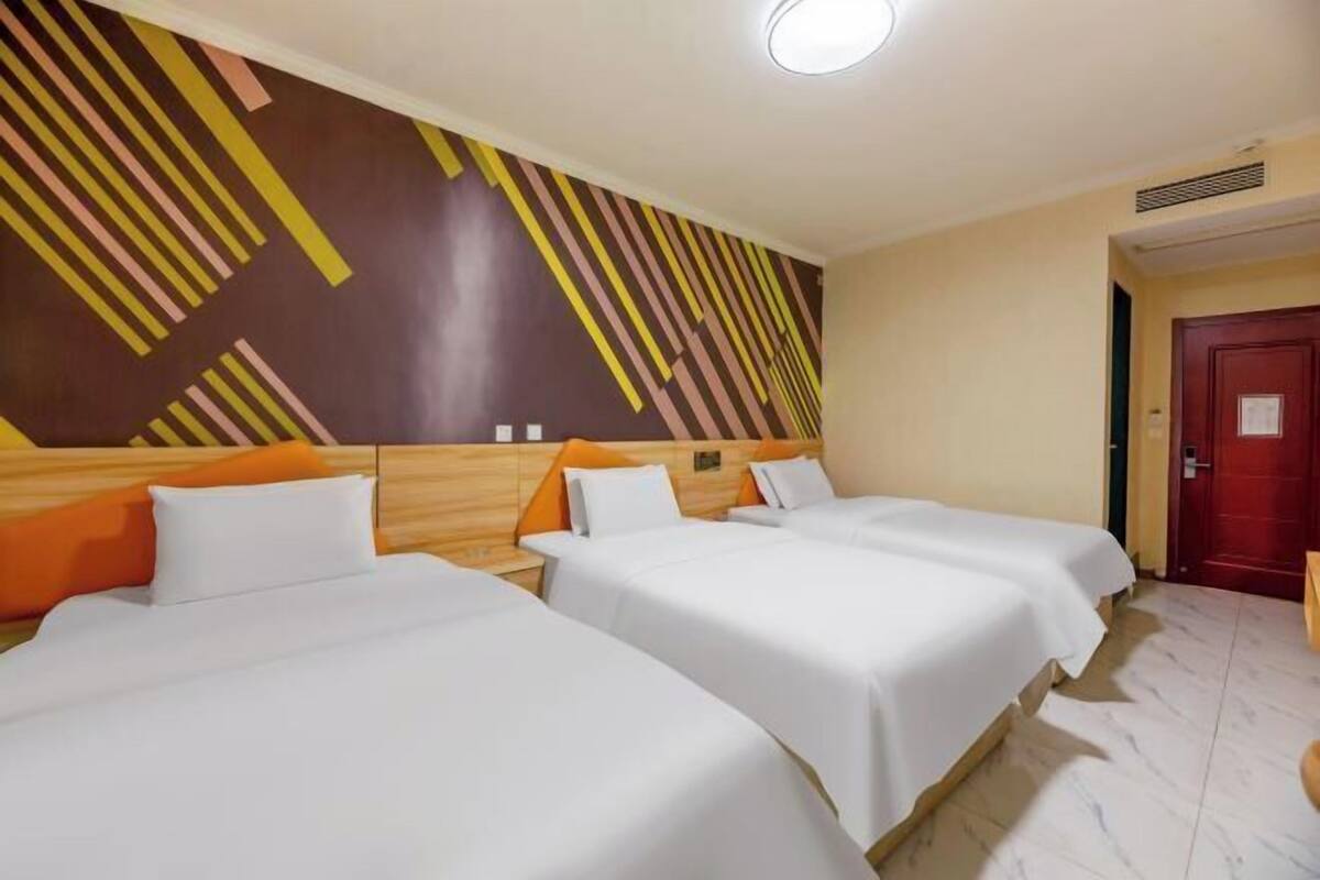 Une chambre d’hôtel avec trois lits, chacun doté de draps blancs et d’oreillers orange. La chambre présente un design moderne, avec un mur orné de motifs géométriques et une porte rouge.